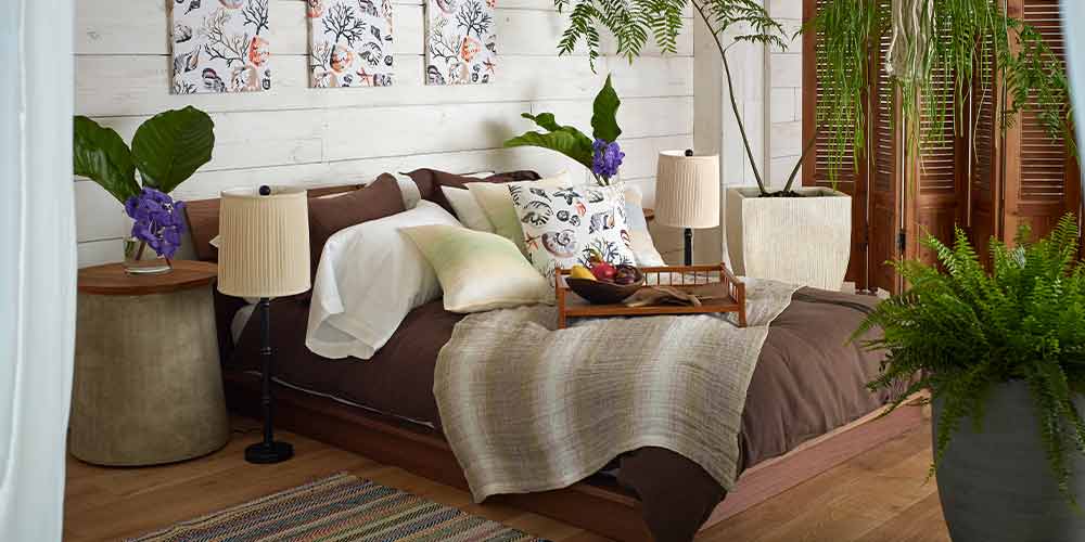 アジアンリゾート風ベッドルーム　観葉植物が置かれた寝室にブラウン・ベージュ色の寝装品でベッドメーキングされたベッドの上にフルーツの乗った木のトレー　ベッド両サイドにテーブルスタンドが床置きされている