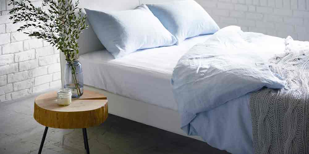 ベッドルームイメージ画像　ライトブルー・ホワイト色の寝装品でコーディネートされたベッドの左脇にサイドテーブルが置かれている（上には透明の花瓶に葉のついた枝が二本挿してある）